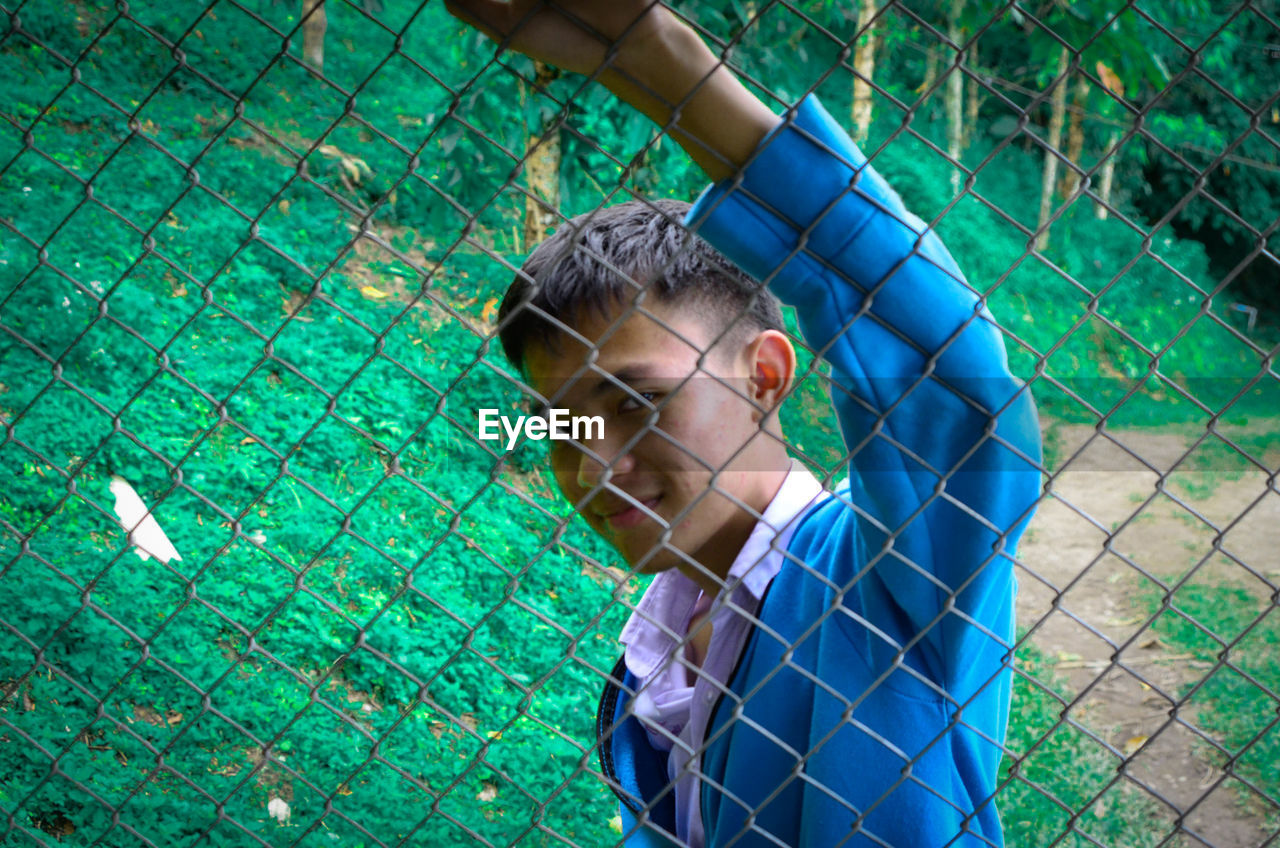 PORTRAIT OF BOY WEARING CHAINLINK FENCE AGAINST GREEN WIRE