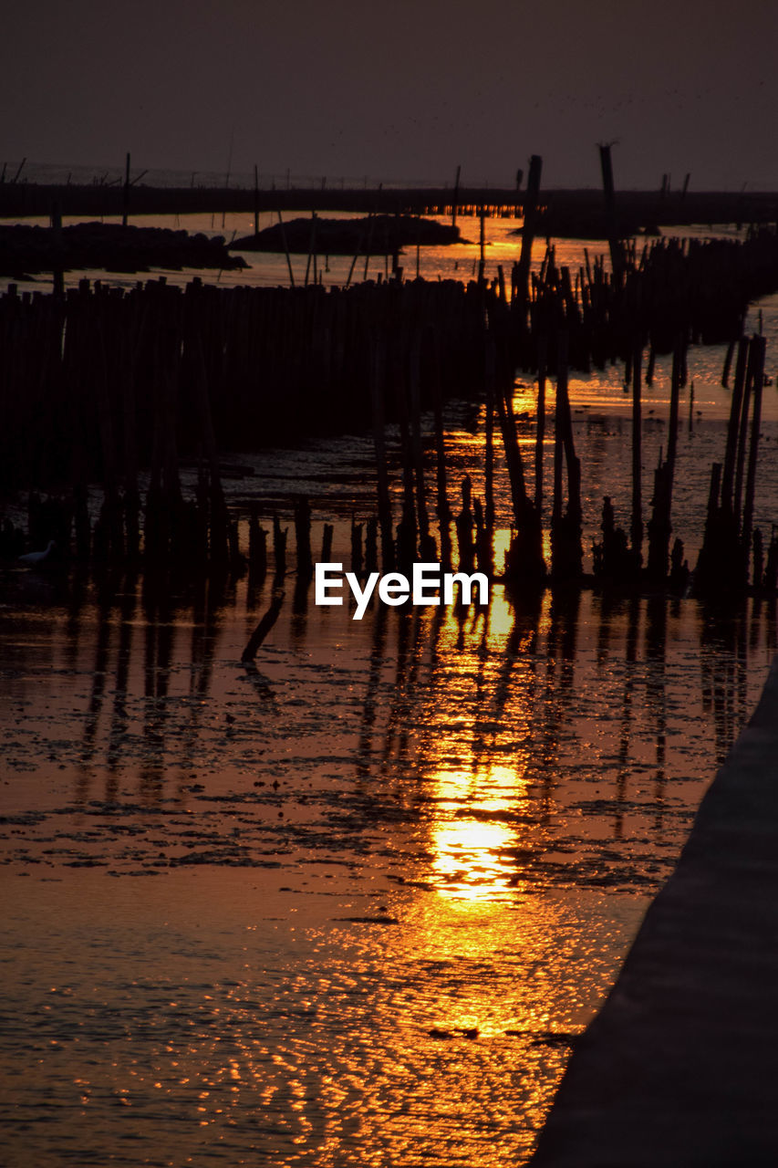 SILHOUETTE WOODEN POSTS IN LAKE AGAINST SKY AT NIGHT