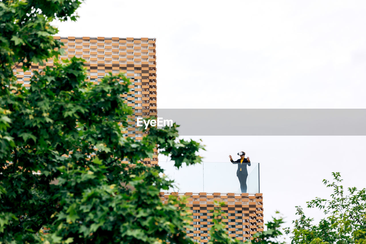 Low angle of female in stylish outfit using vr goggles while standing on terrace of house near glass railings under cloudy sky in daylight near green trees and plants