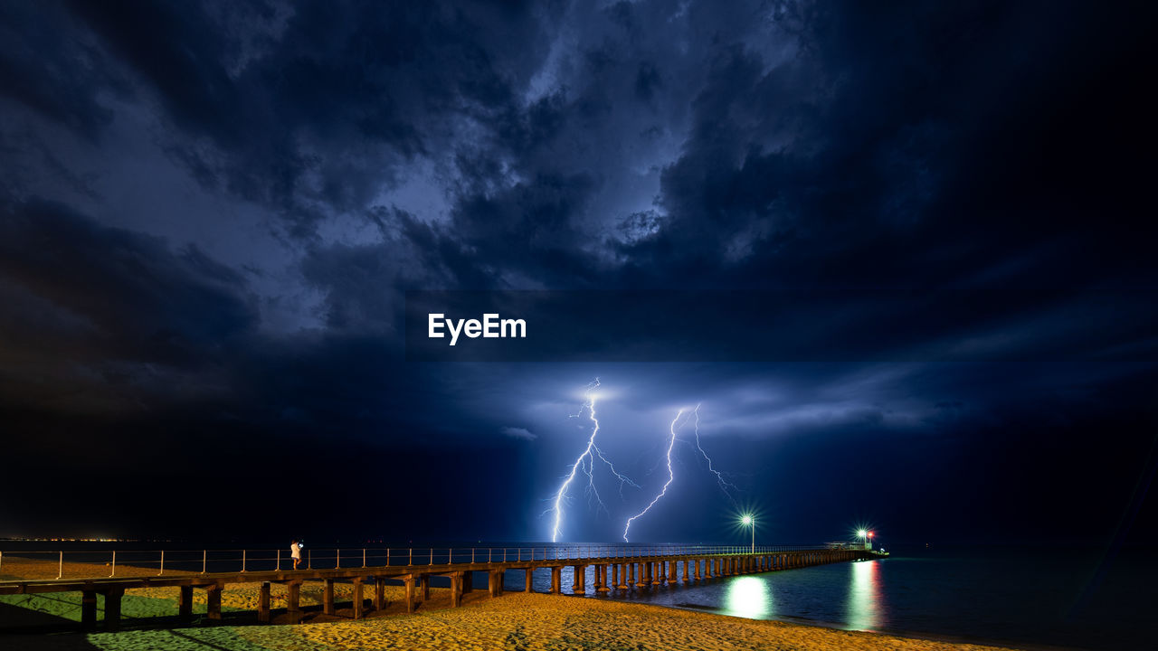 Image of a lightning strike with a pier in the foreground and a person taking a photo of the storm.