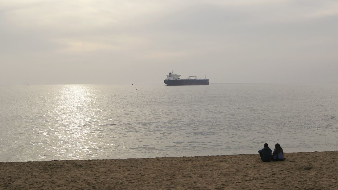 Couple sitting on beach looking at ship against clear sky
