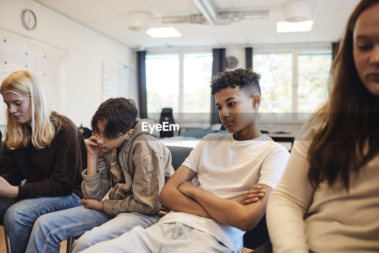 Thoughtful teenage boy sitting with arms crossed amidst friends in classroom during group therapy