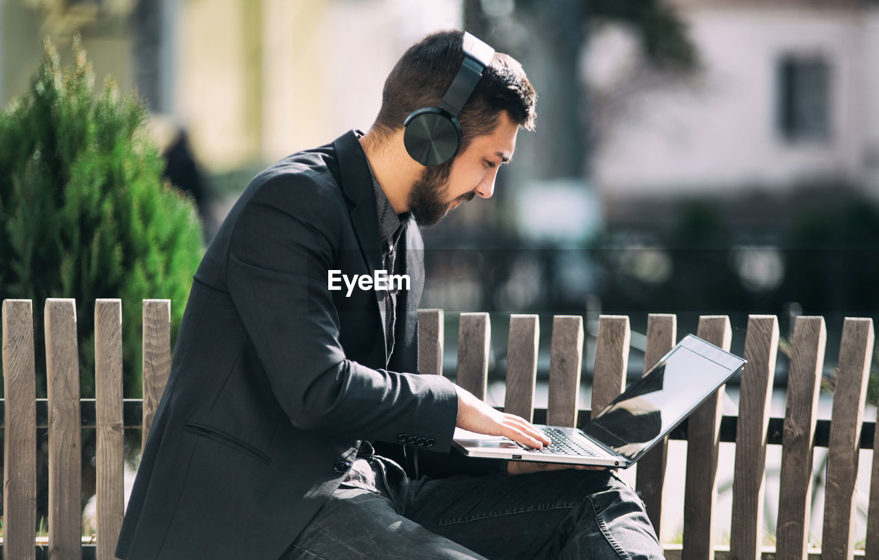 Side view of businessman using laptop while listening music on bench