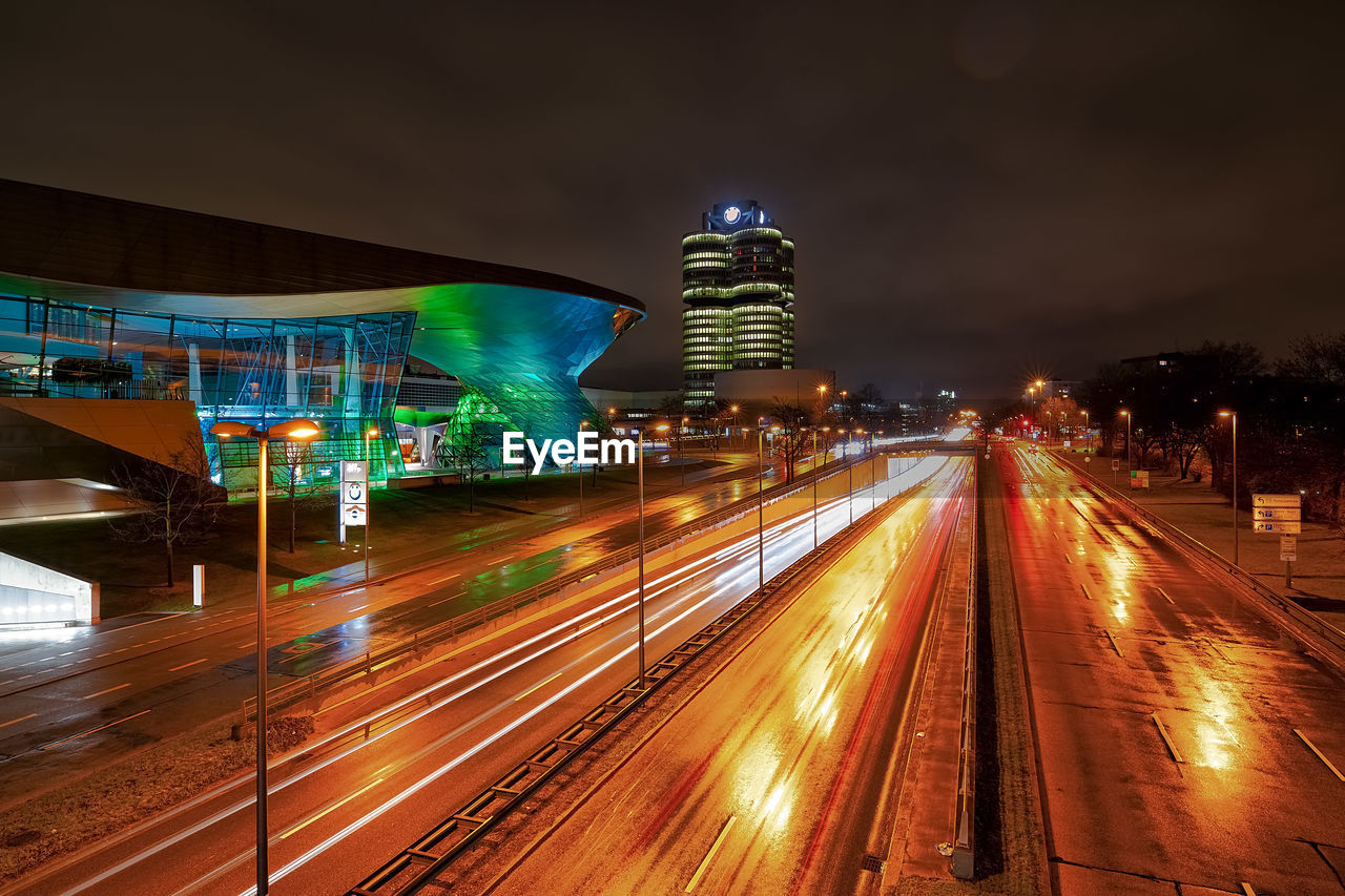 LIGHT TRAILS ON ROAD BY ILLUMINATED BUILDINGS AT NIGHT