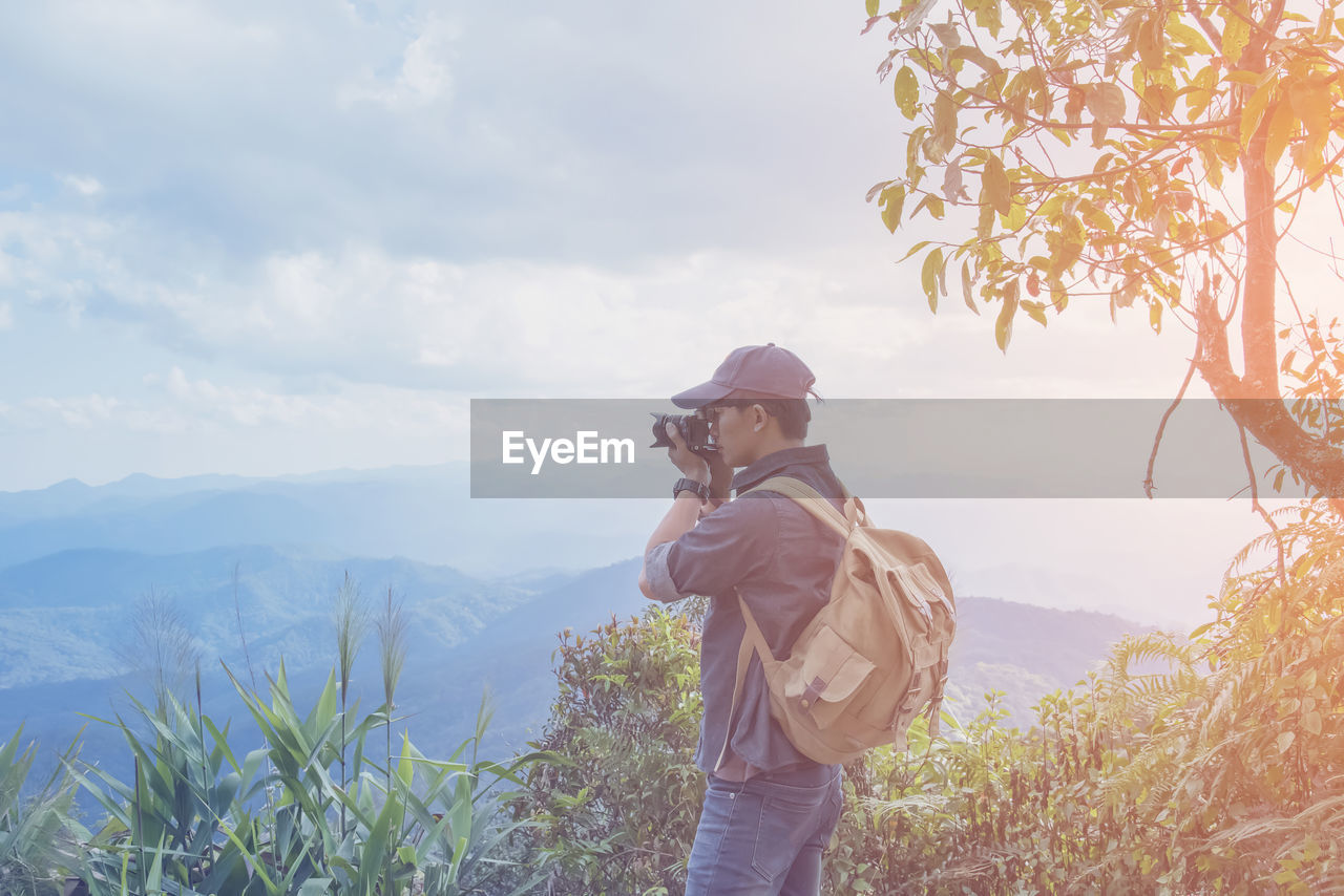 Side view of man photographing while standing on mountain against sky