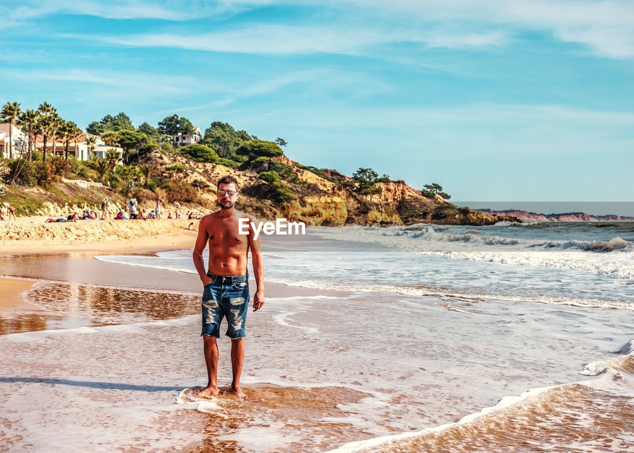 FULL LENGTH REAR VIEW OF SHIRTLESS MAN STANDING ON BEACH