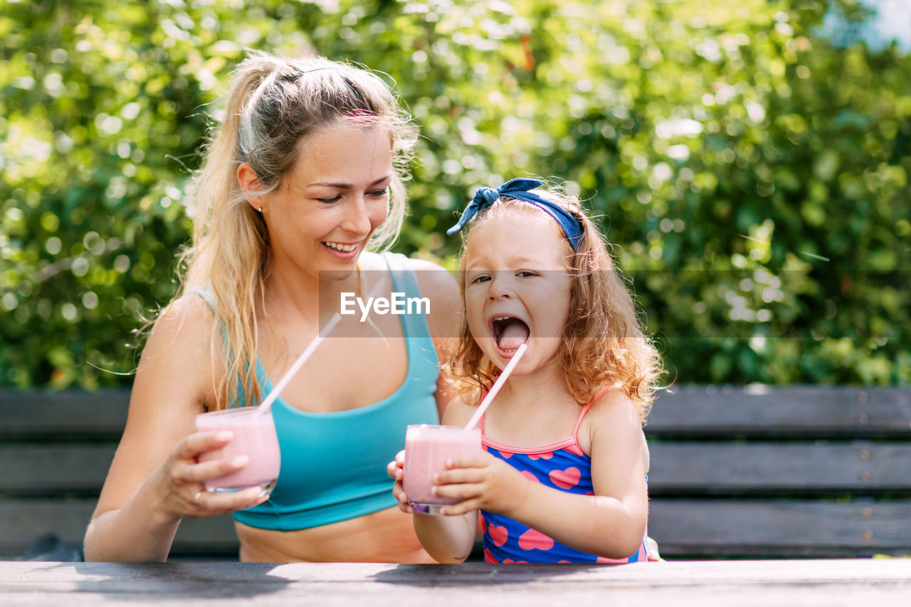 A beautiful girl and her little daughter drink a refreshing smoothie while sitting on a bench 