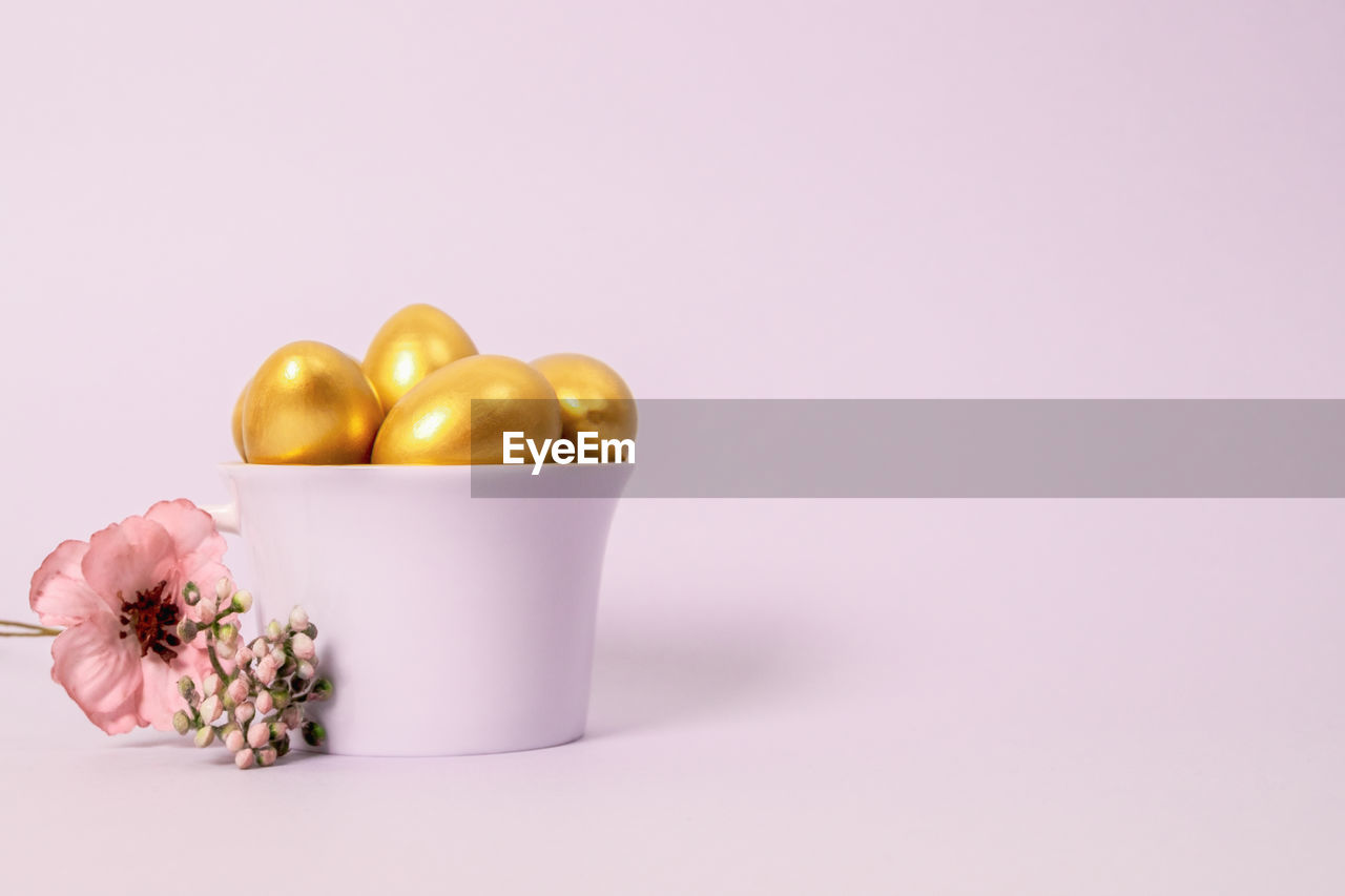Easter composition of golden decorative eggs in a porcelain cup and a flower on a pink background. 