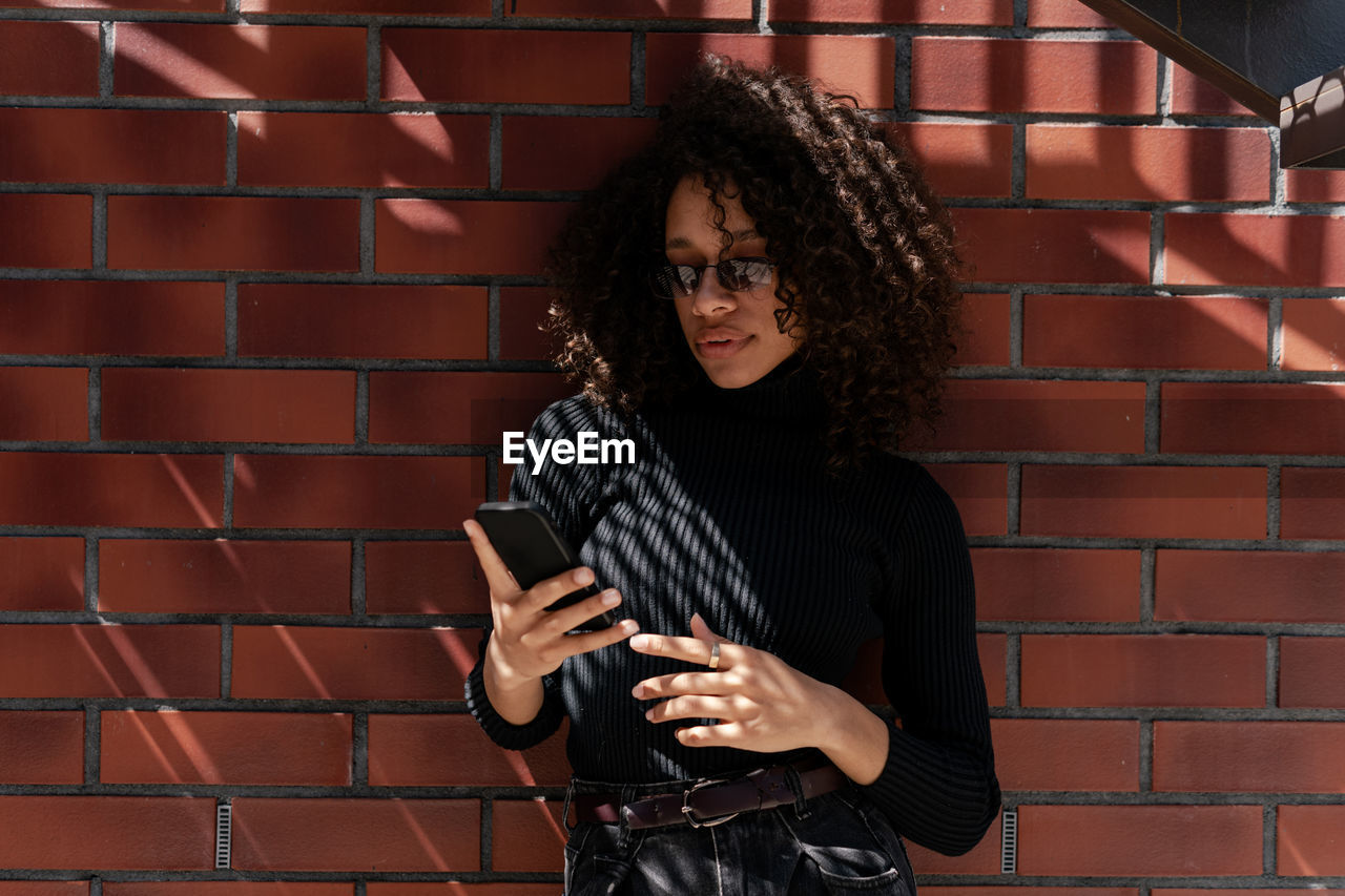 Young creative woman using her cellphone while standing against a brick wall