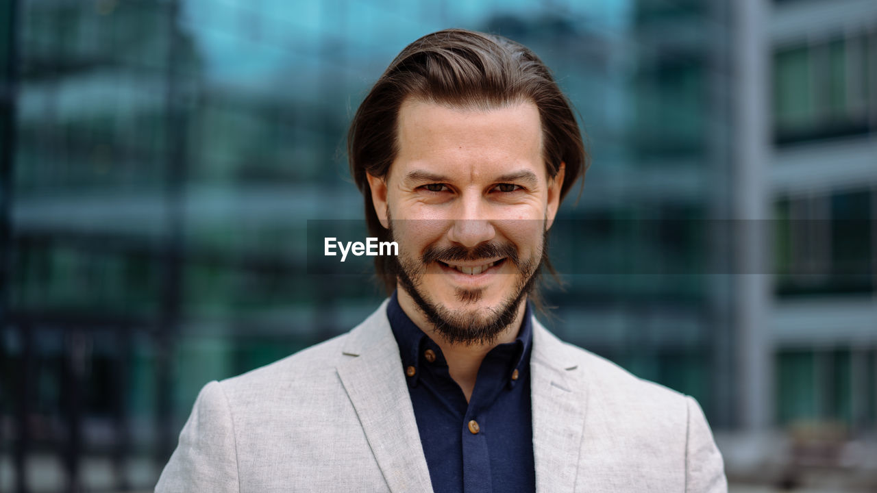 Smiling positive bearded professional businessman in suit looking at camera while standing on street