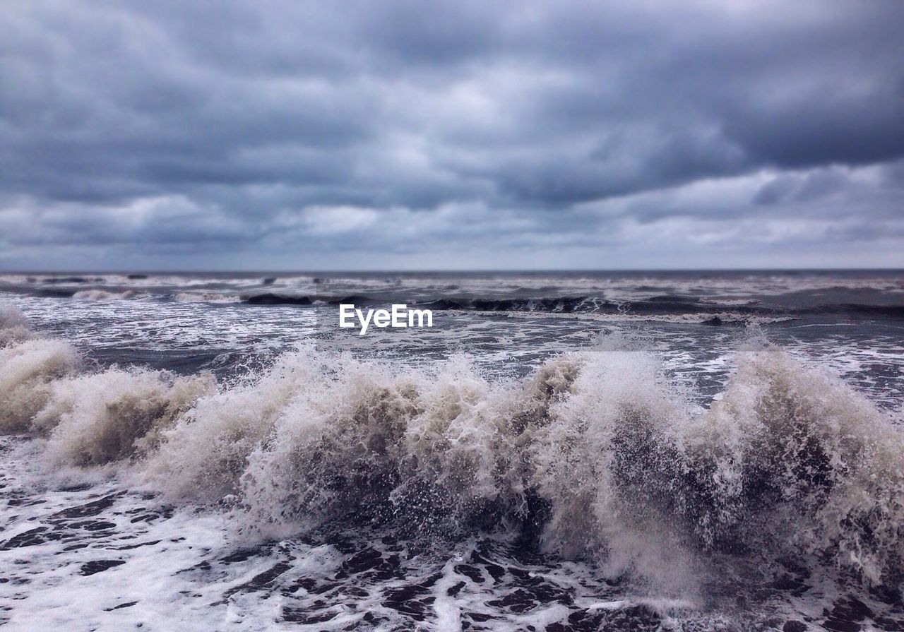 Waves rushing towards at beach against cloudy sky