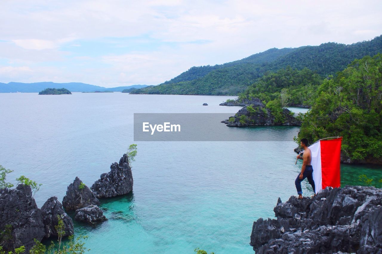 High angle view of shirtless man standing with indonesian flag on cliff against sea