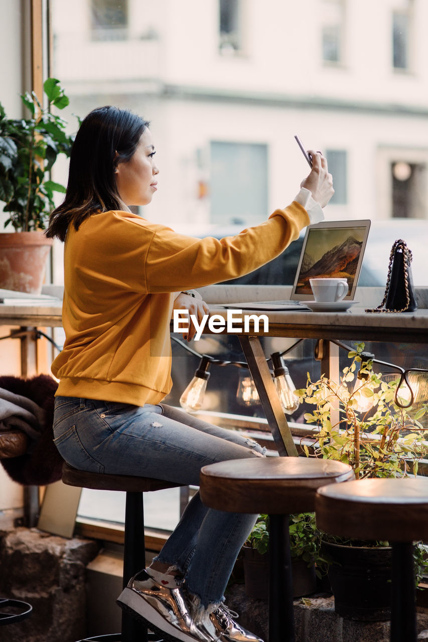 Full length side view of female blogger taking selfie sitting at cafe