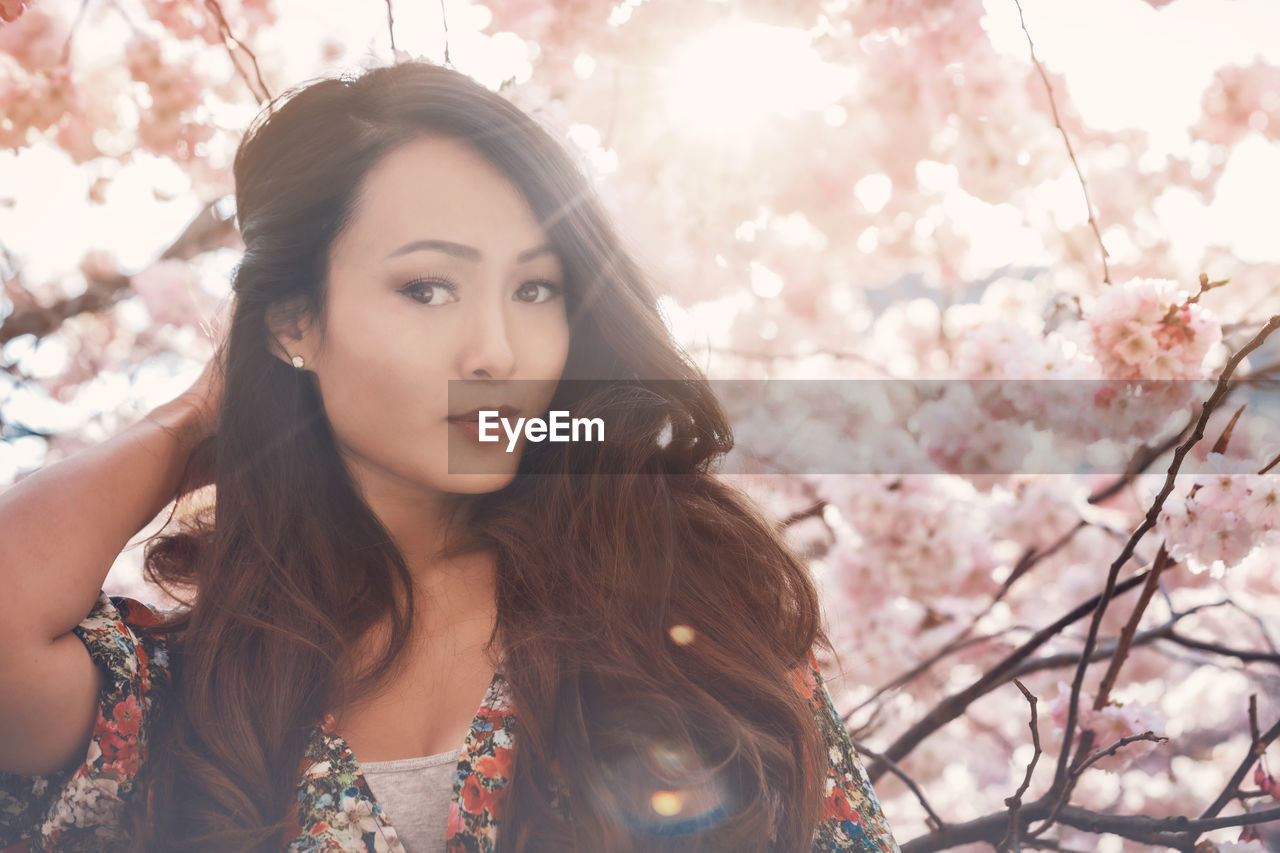 spring, plant, young adult, one person, tree, women, flower, adult, portrait, flowering plant, blossom, female, springtime, nature, long hair, hairstyle, beauty in nature, cherry blossom, sunlight, brown hair, fragility, freshness, lens flare, back lit, looking, front view, fashion, sky, sunbeam, outdoors, looking at camera, headshot, photo shoot, lifestyles, clothing, branch, tranquility, day, pink, cherry tree, low angle view, skin, contemplation, emotion, black hair, waist up, leisure activity, casual clothing, relaxation, human face, person, human eye, happiness, standing, focus on foreground, sunset, copy space, human hair