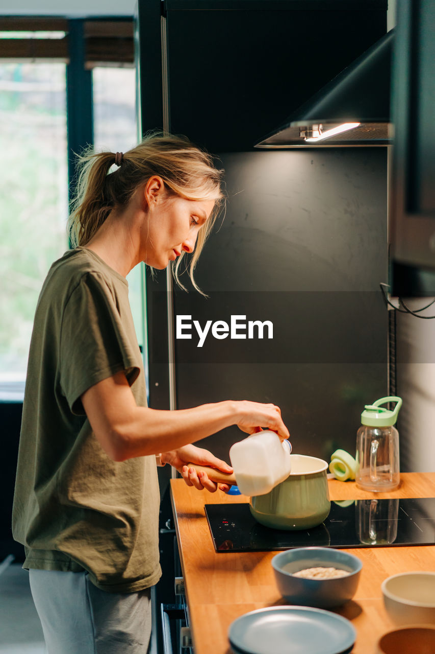 Young housewife woman in the kitchen prepares breakfast milk porridge on the stove.