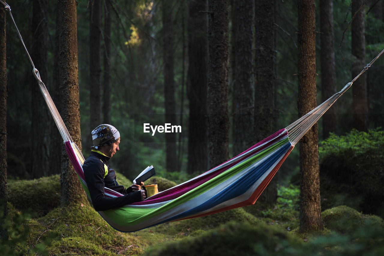 Man reading book while sitting in hammock at forest