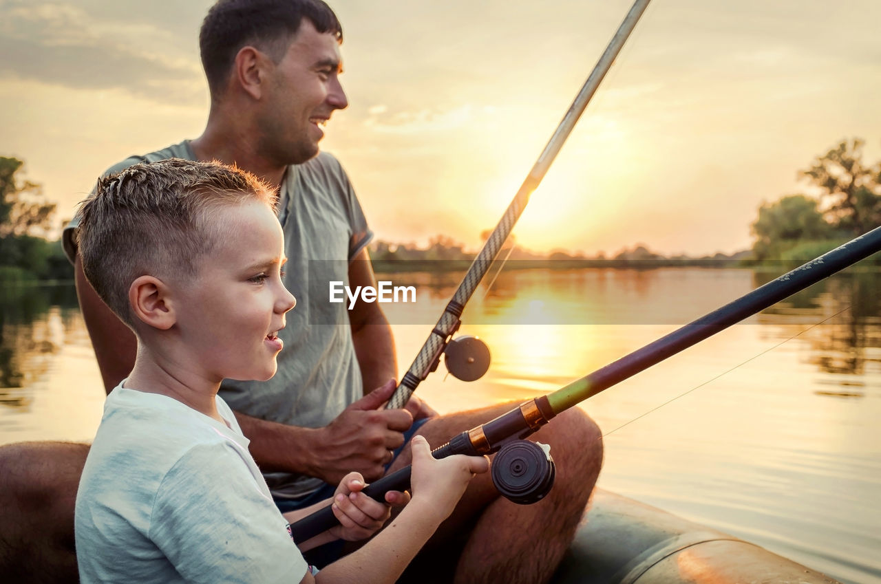 Happy father and son together fishing from a boat at sunset time, summer day, beautiful sky on lake.