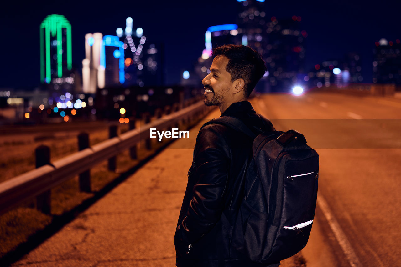 Side view of fashionable hispanic man in casual black leather jacket with backpack looking away with night city on blurred background