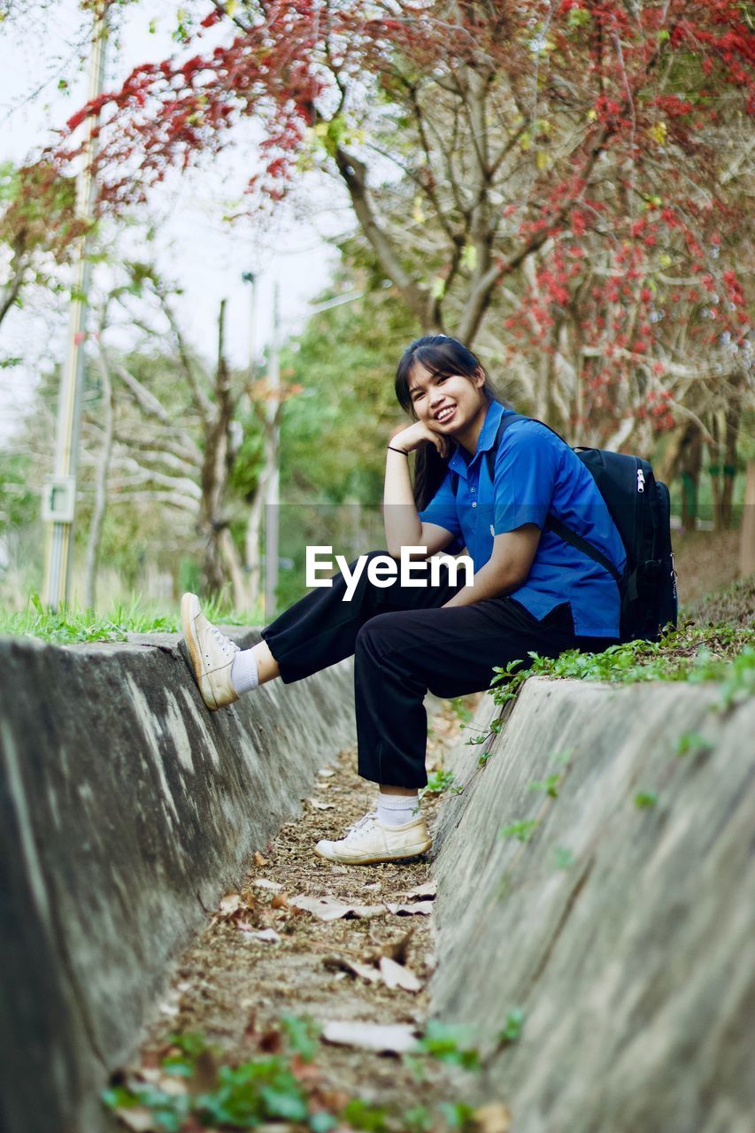 Portrait of smiling young woman with backpack sitting against plants in park