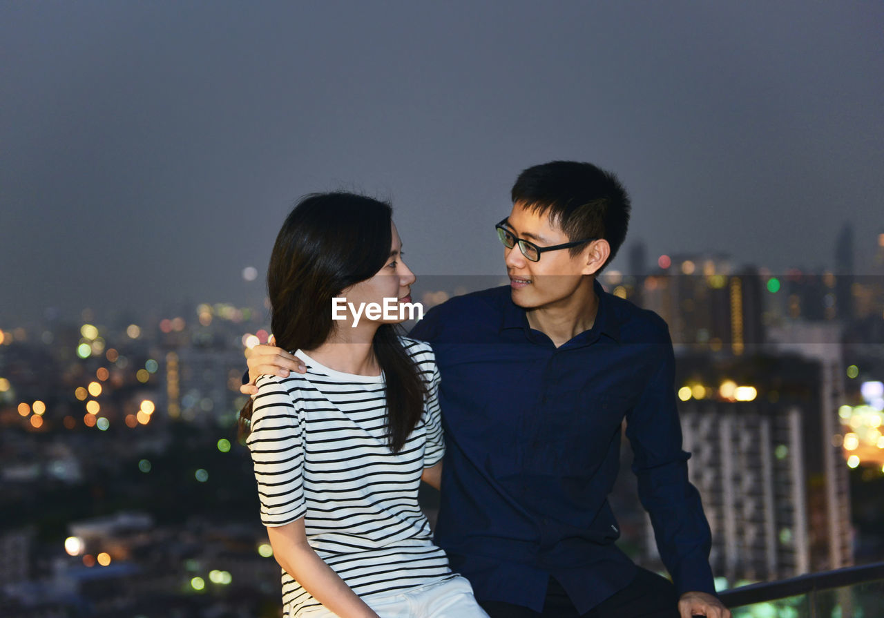 Couple sitting on railing against illuminated city at dusk
