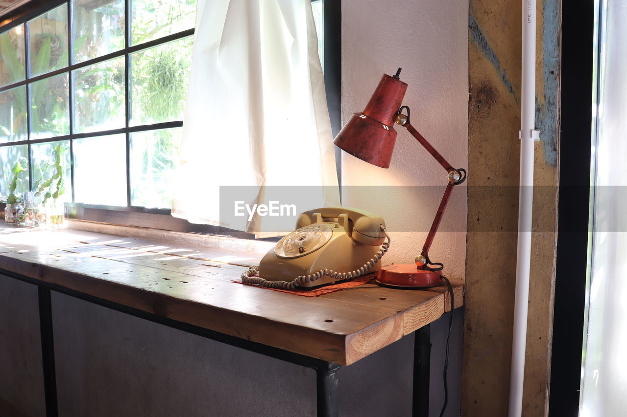 An old rotary phone and an electric lamp on a wooden table near the glass window and fabric curtain