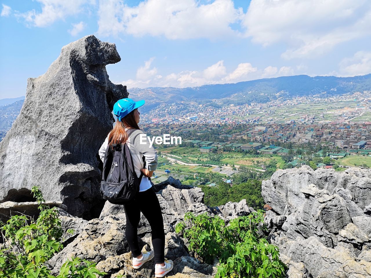 WOMAN STANDING ON ROCK LOOKING AT MOUNTAINS AGAINST SKY