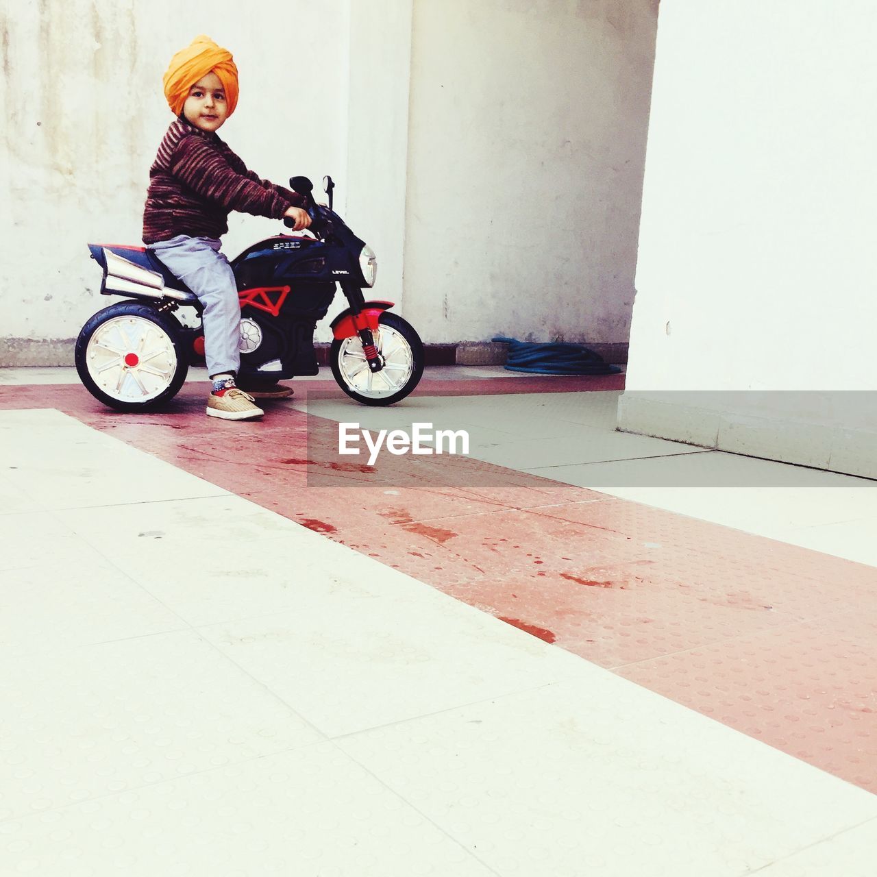Side view of cute boy with turban sitting on toy motorcycle outside house
