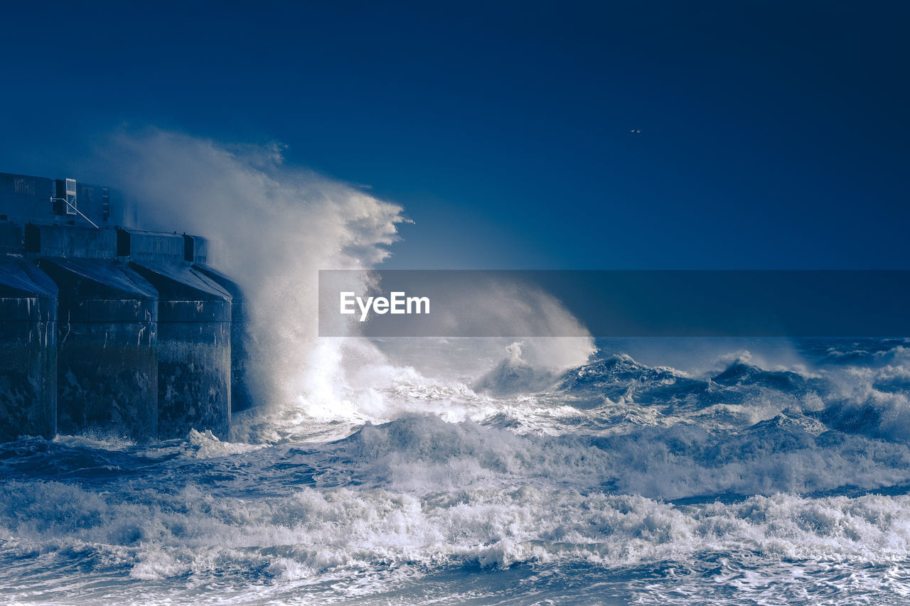 Sea waves splashing on bridge against clear blue sky at night