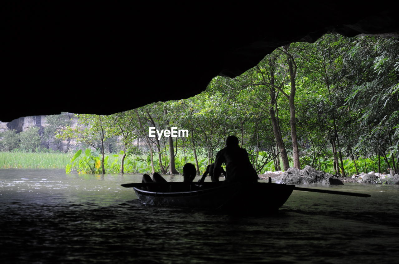 Rear view of silhouette men in boat sailing on river below cave