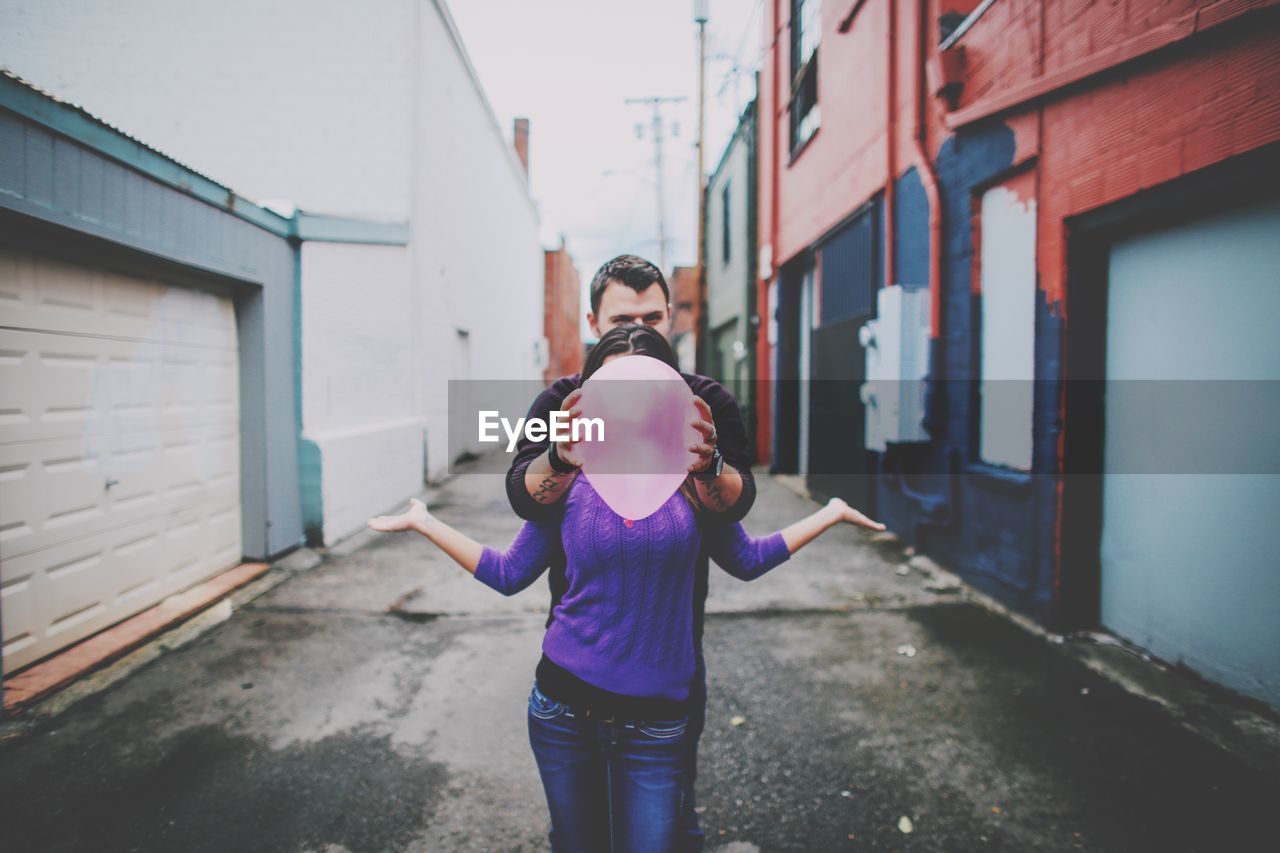 Man holding balloon over woman face while standing in alley