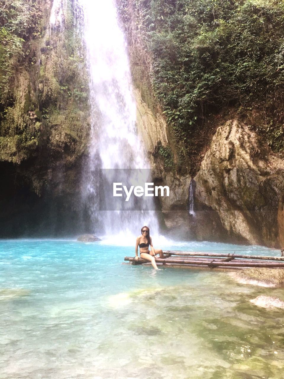 Woman in bikini sitting on wooden raft against waterfall