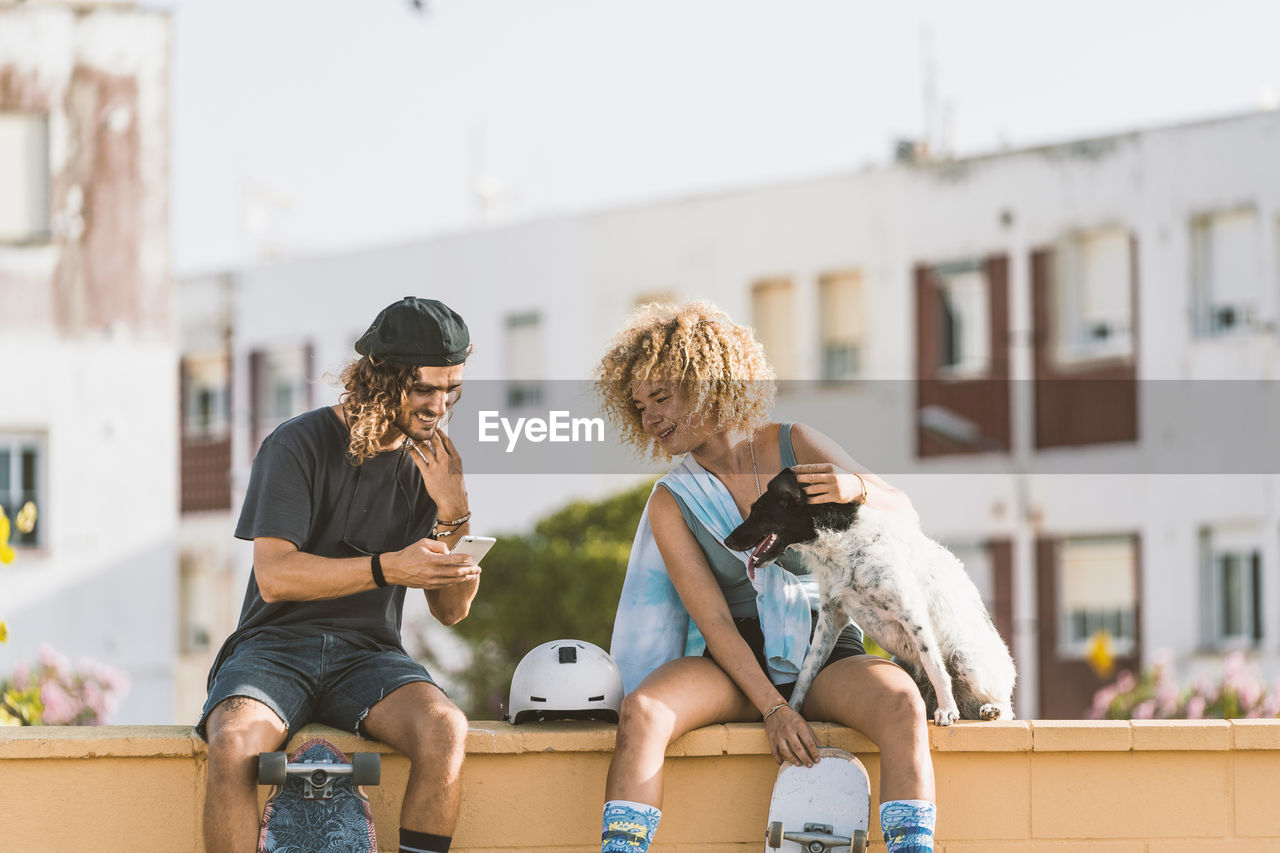 Friends using mobile phone while sitting with dog on retaining wall in city