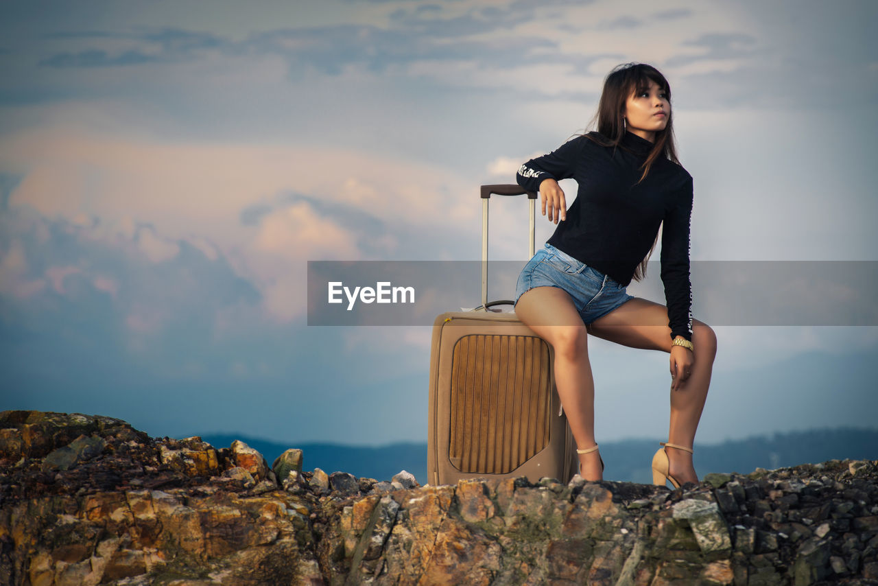 Young woman sitting on suitcase on cliff against cloudy sky