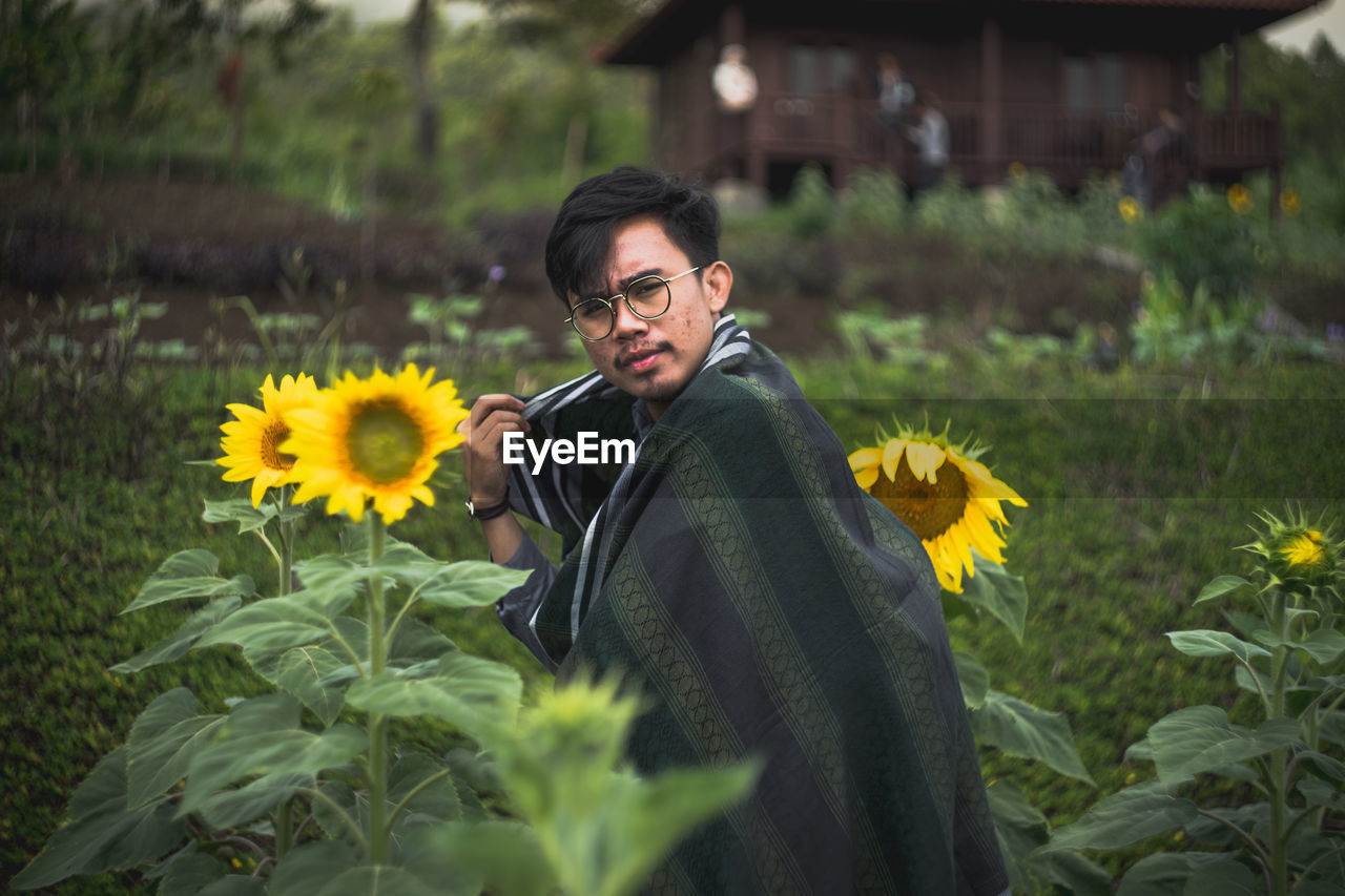 Portrait of young man standing amidst sunflowers blooming on field