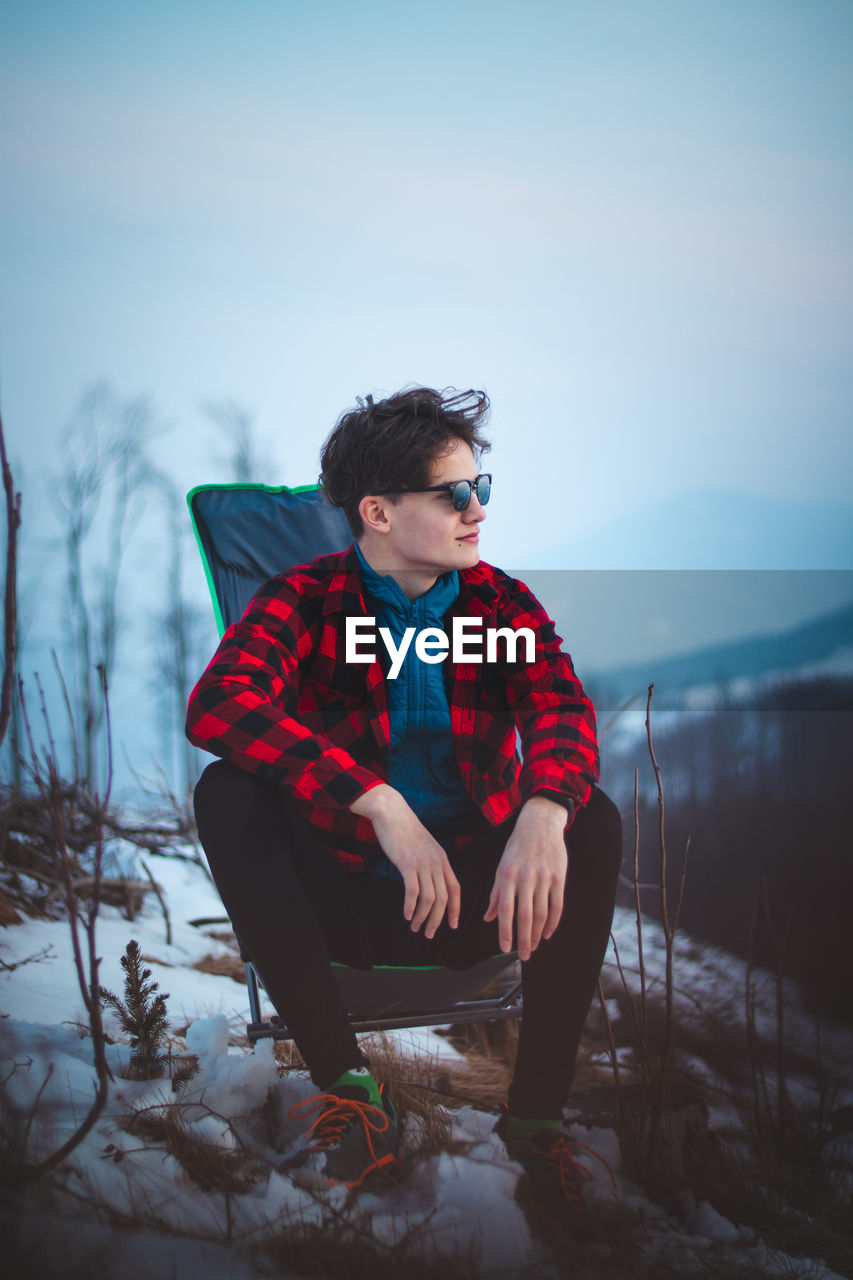 Handsome teenager aged 15-20 in a plaid red-black shirt and sunglasses on a chair by the tent. 