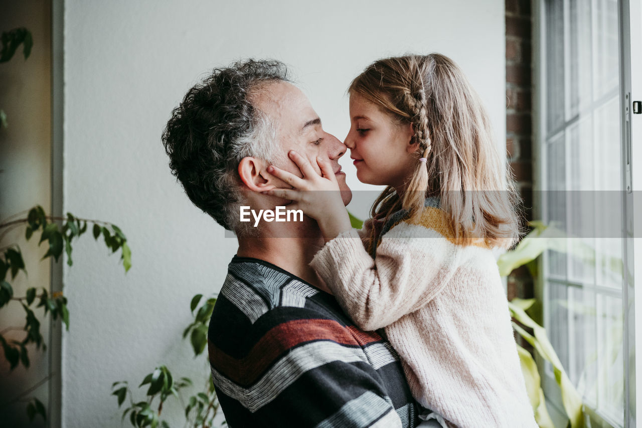 Daughter rubbing nose with father while standing by window at home