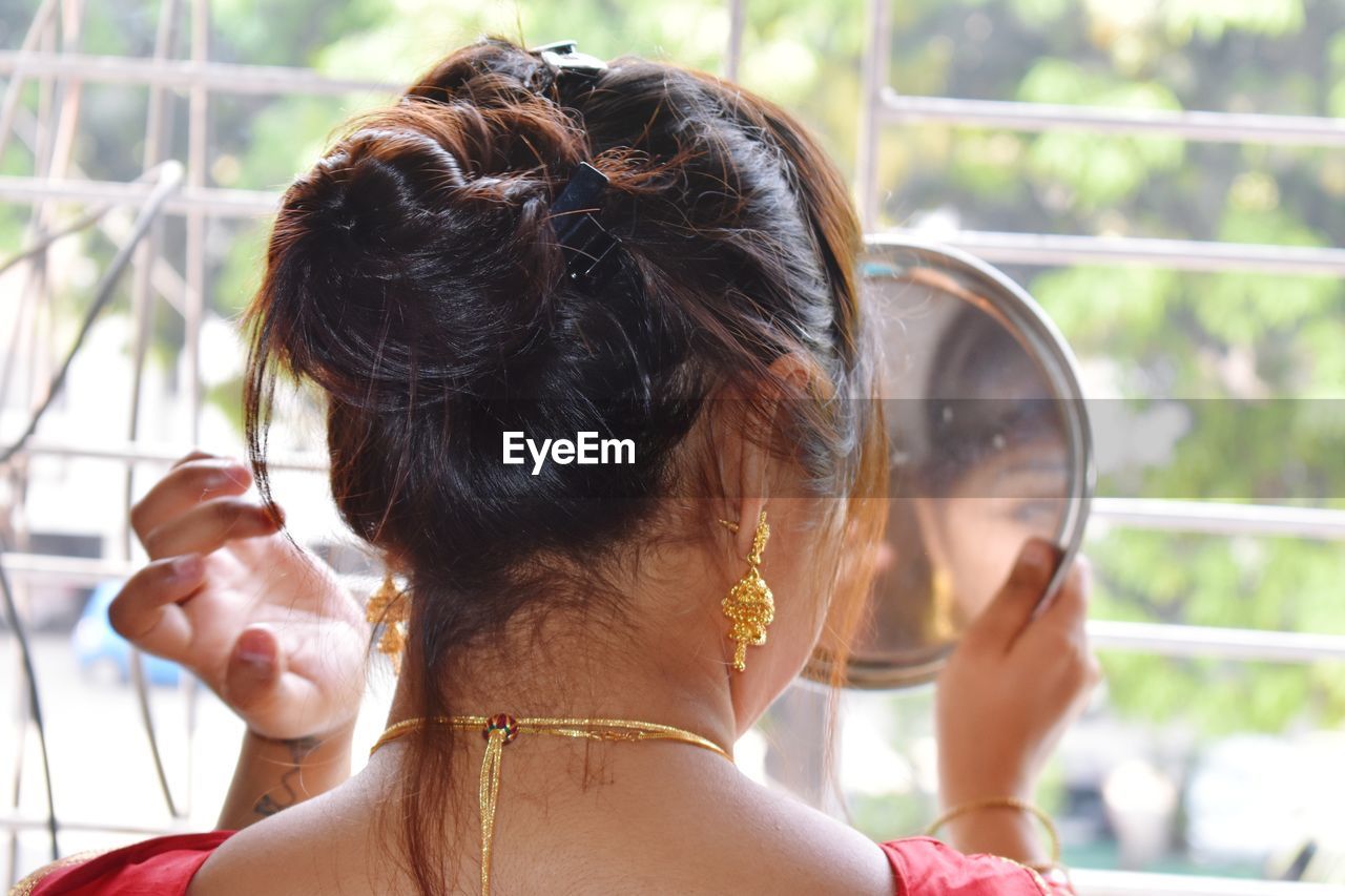 Rear view of woman looking in hand mirror against window