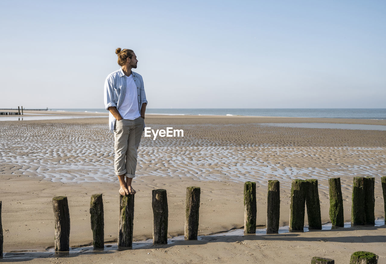 Young man looking at view while standing on wooden post at beach against clear sky