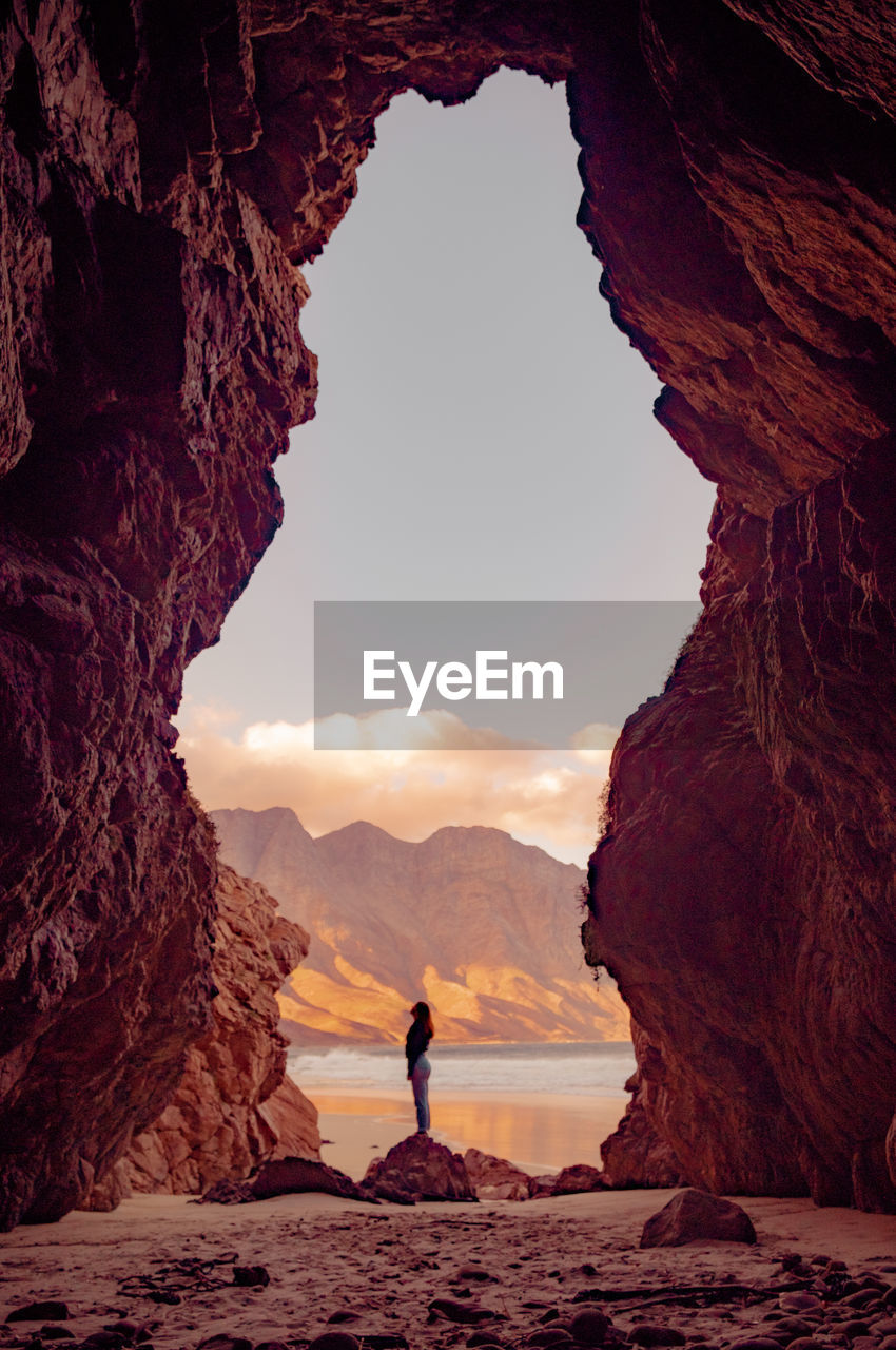 Woman standing under rock formation at beach against sky