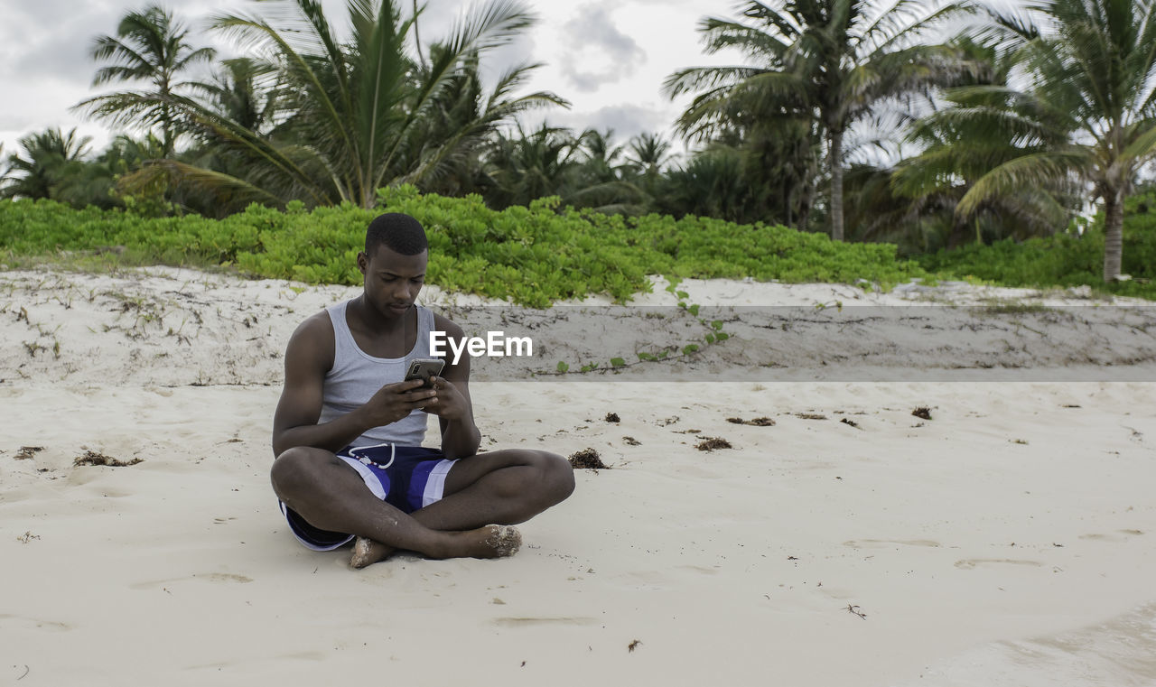 From the front, black traveler sitting on the sand and browsing social networks on cell phone