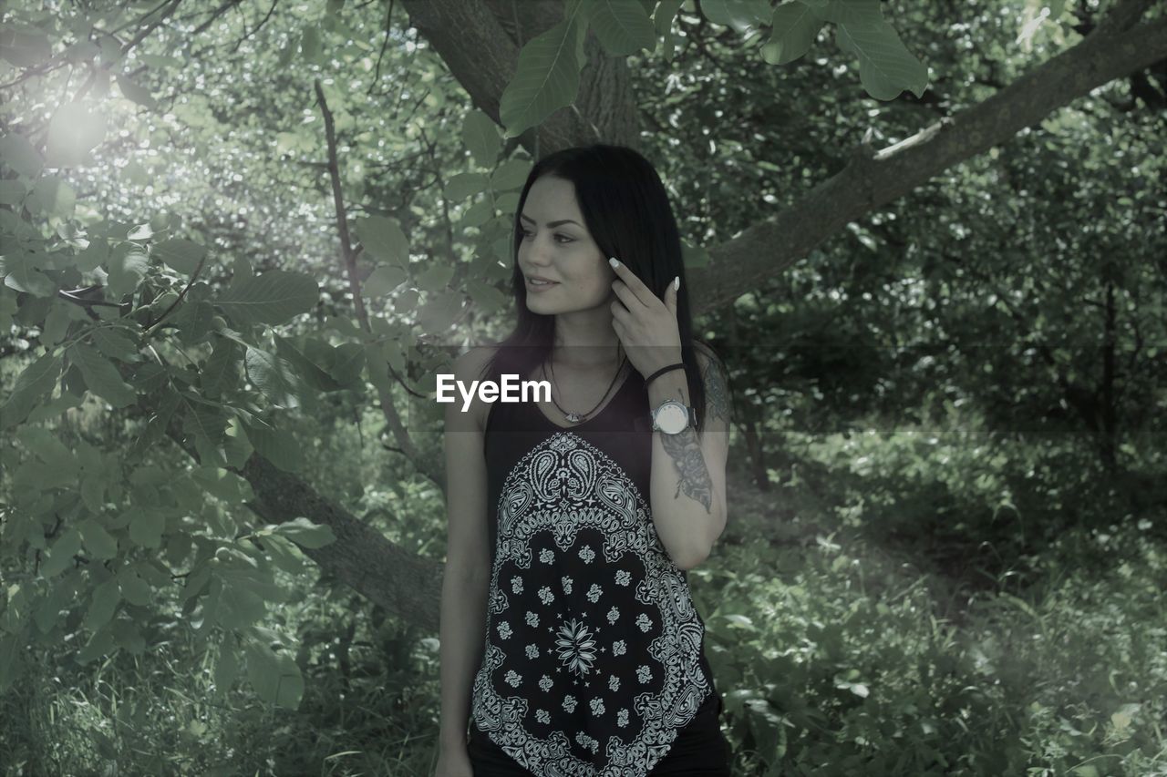 Beautiful smiling young woman with hand in hair standing against trees