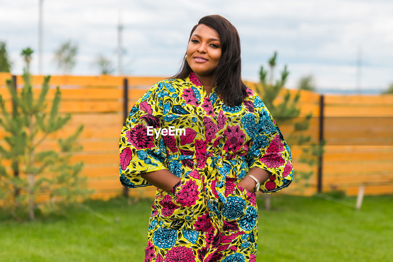 one person, spring, women, smiling, adult, yellow, happiness, standing, dress, young adult, portrait, nature, emotion, clothing, flower, lifestyles, multi colored, grass, cheerful, floral pattern, hairstyle, front view, pattern, looking at camera, female, plant, focus on foreground, outdoors, fashion, three quarter length, photo shoot, sky, day, brown hair, leisure activity, architecture, teeth, waist up, smile, casual clothing, person, enjoyment, long hair, green, sleeve, copy space, human face