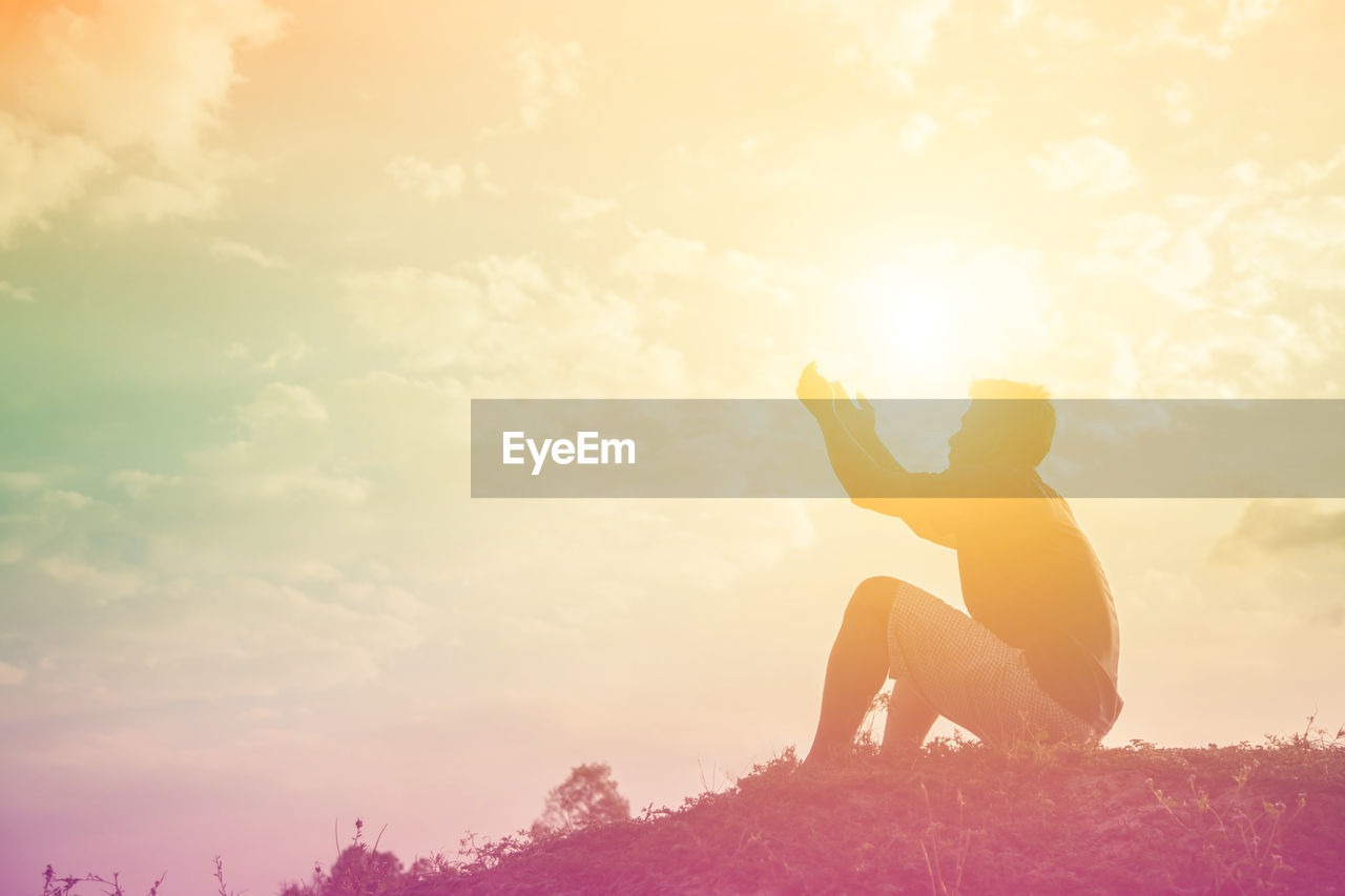 Side view of man gesturing while sitting on land against sky during sunset