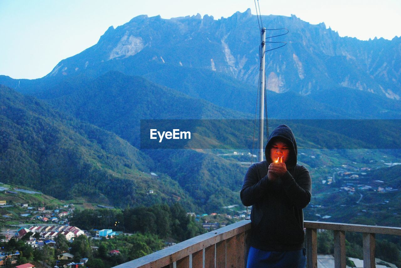 Man with hooded shirt igniting cigarette by railing against mountains