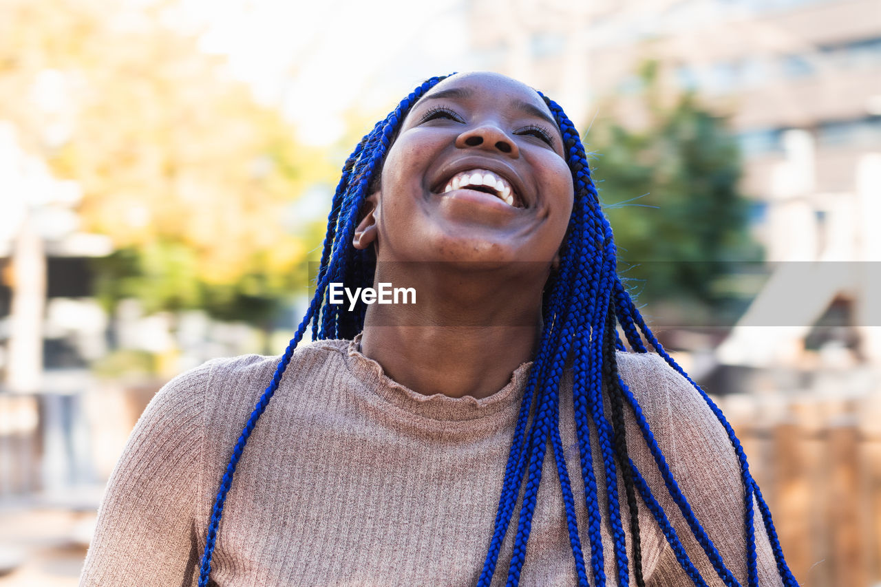 Charming african american female with blue braids sincerely laughing with closed eyes while standing on street