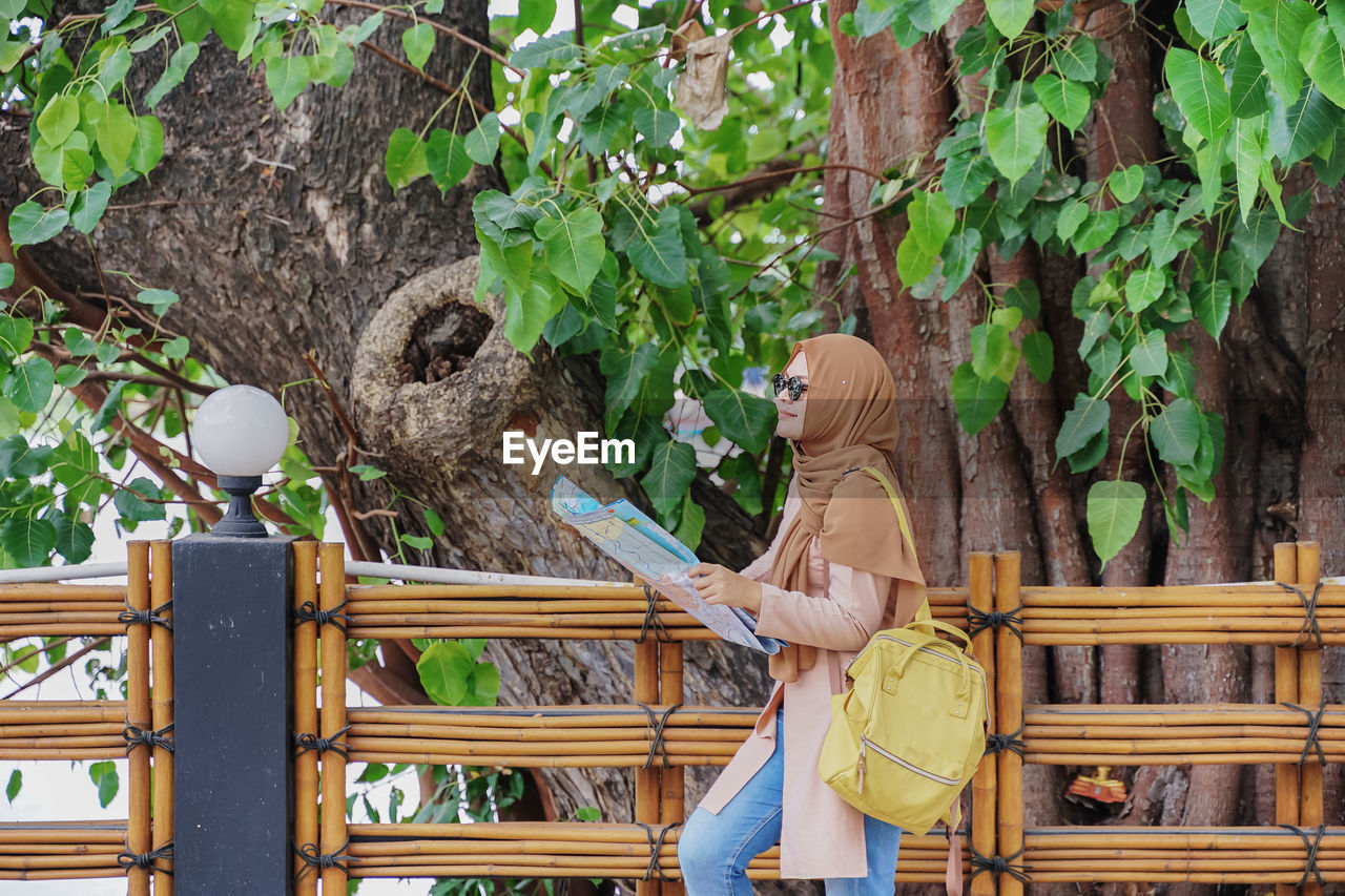 Woman reading map while standing by railing against trees