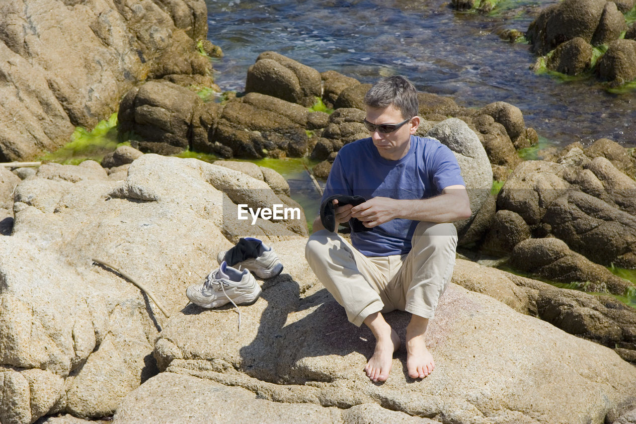 High angle view of mature man sitting on rock formation by stream during sunny day