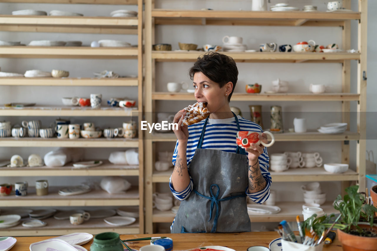 Relaxed woman in apron drink coffee from handmade ceramic cup and has snack in pottery studio