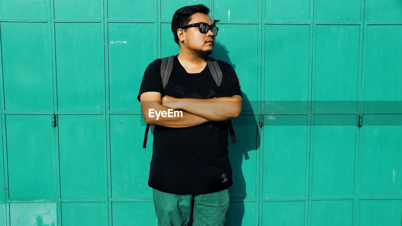 Young man standing with arms crossed against green wall on sunny day