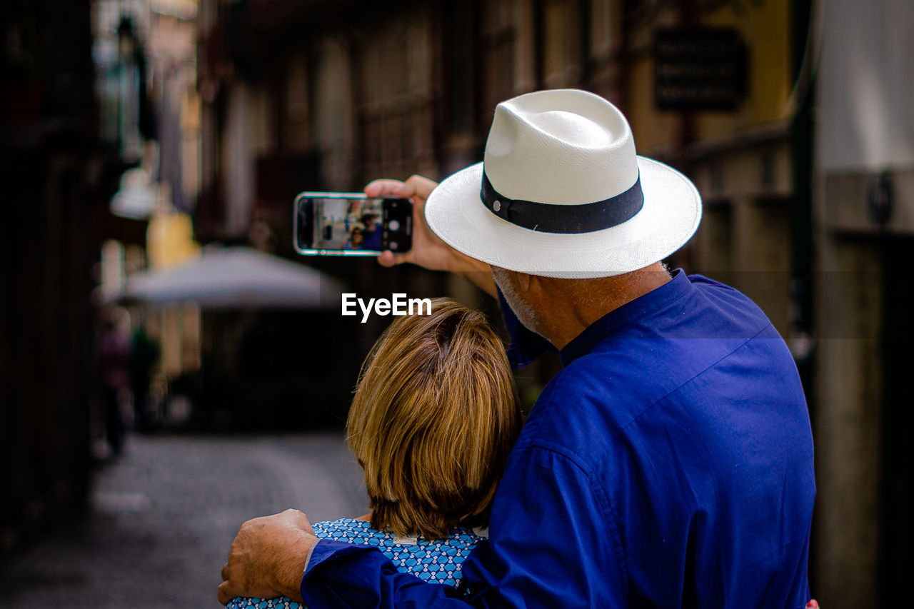 adult, hat, two people, technology, men, person, rear view, clothing, city, fashion accessory, women, photographing, wireless technology, communication, togetherness, cowboy hat, architecture, portable information device, smartphone, blue, activity, fedora, sun hat, street, outdoors, female, lifestyles, portrait, holding